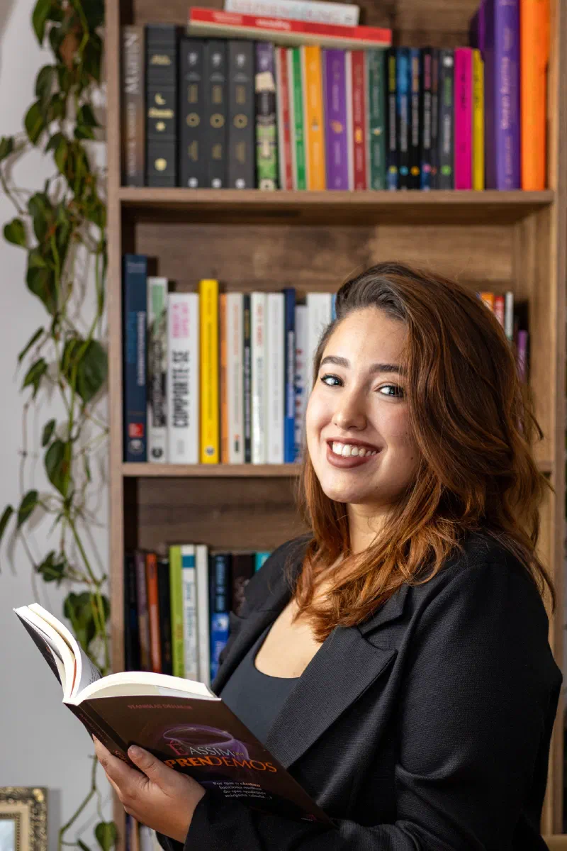 Professora Cássia Santos sorrindo, segurando um livro, em frente a uma estante